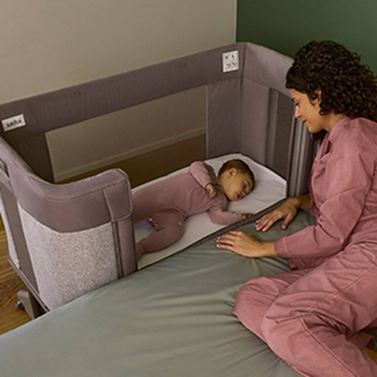 Eine Frau in einem rosa Outfit sitzt auf einem Bett und wacht über ein Baby, das in einem Beistellbettchen neben dem Bett schläft. Der Raum ist sanft beleuchtet und ruhig.