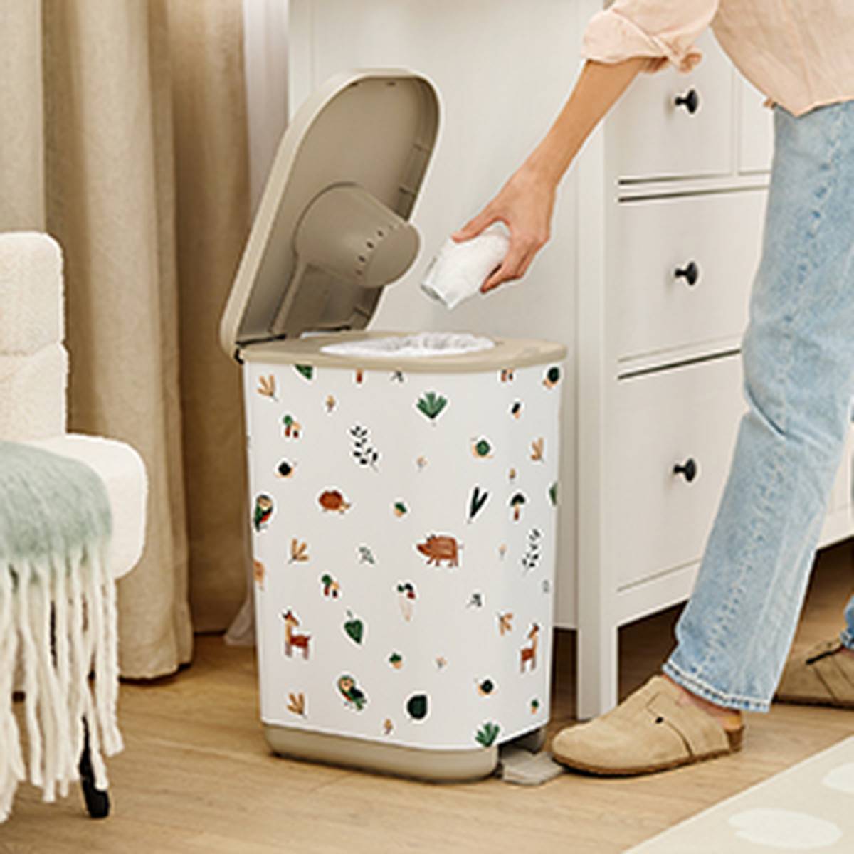 Person placing a diaper in a decorated diaper pail with animal patterns, next to a white dresser and carpeted floor.