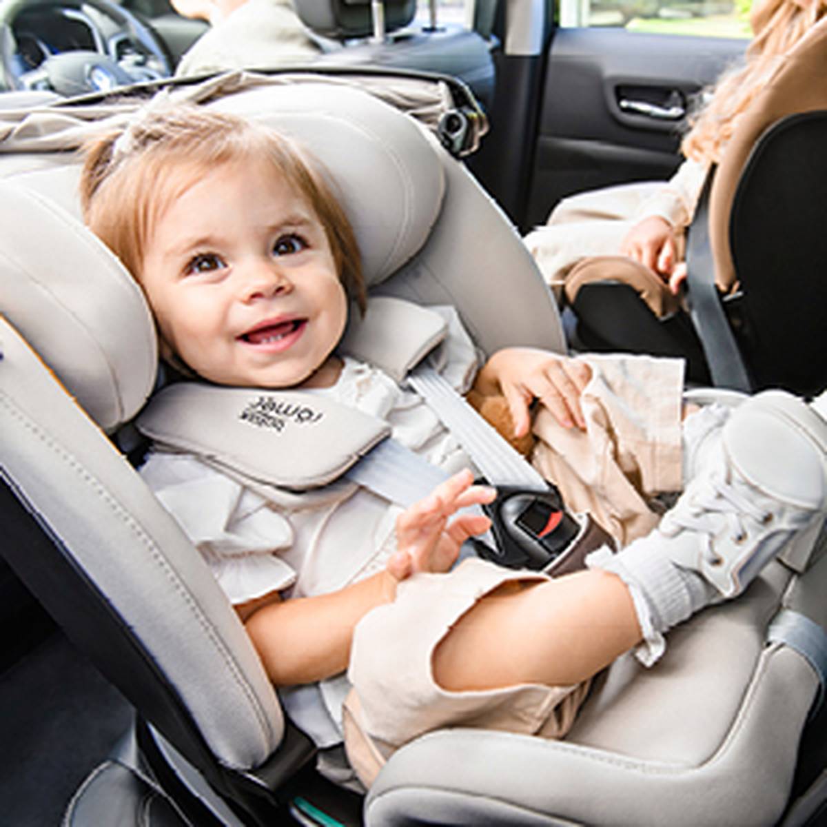 Ein lächelndes Kleinkind sitzt in einem grauen Kindersitz im Inneren eines Fahrzeugs; es trägt ein weißes Outfit sowie Schuhe, und eine erwachsene Person befindet sich auf dem Vordersitz.