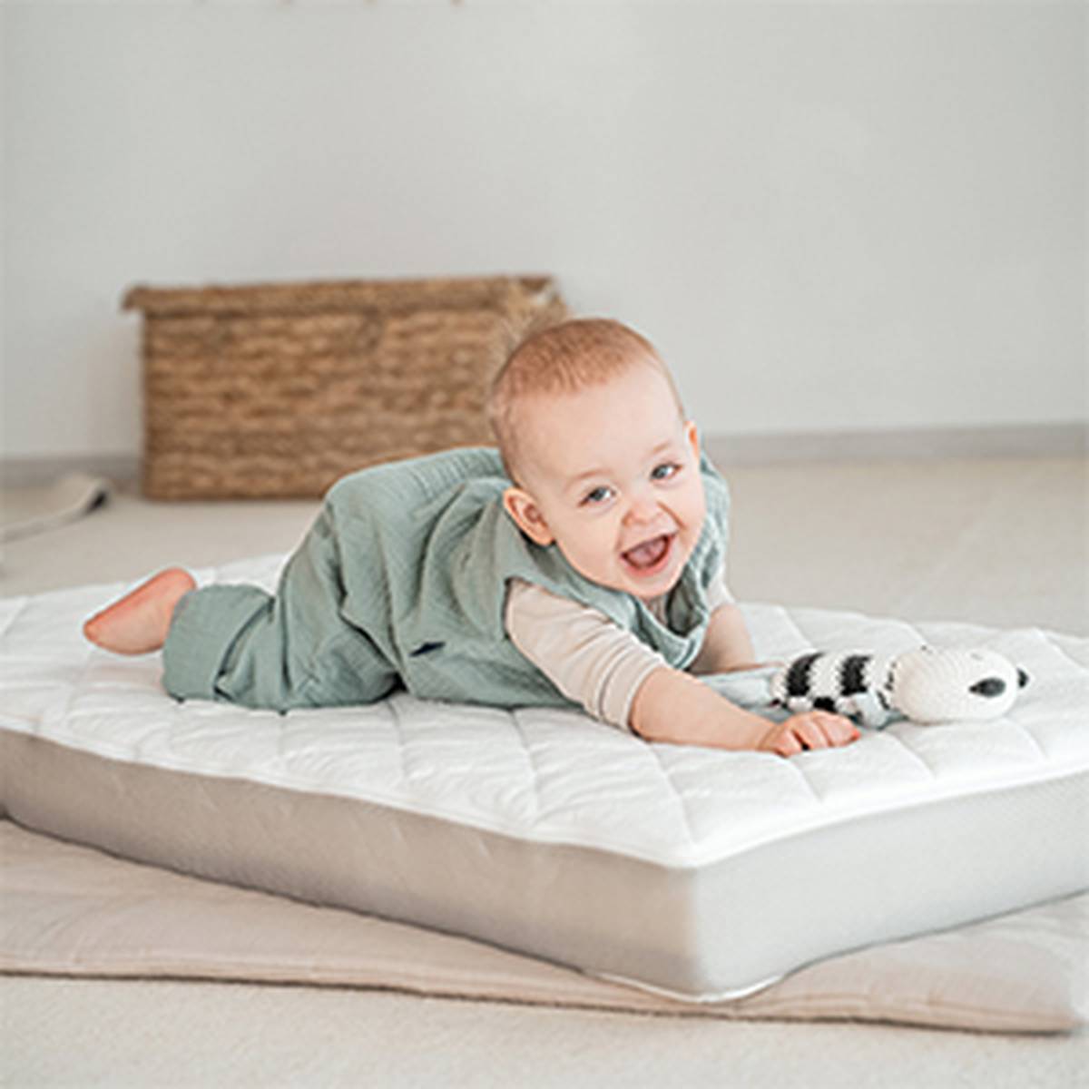 Ein lächelndes Baby in einem grünen Overall liegt auf einer weißen Matratze und hält ein schwarz-weißes Stofftier. Im Hintergrund ist ein Weidenkorb zu sehen.