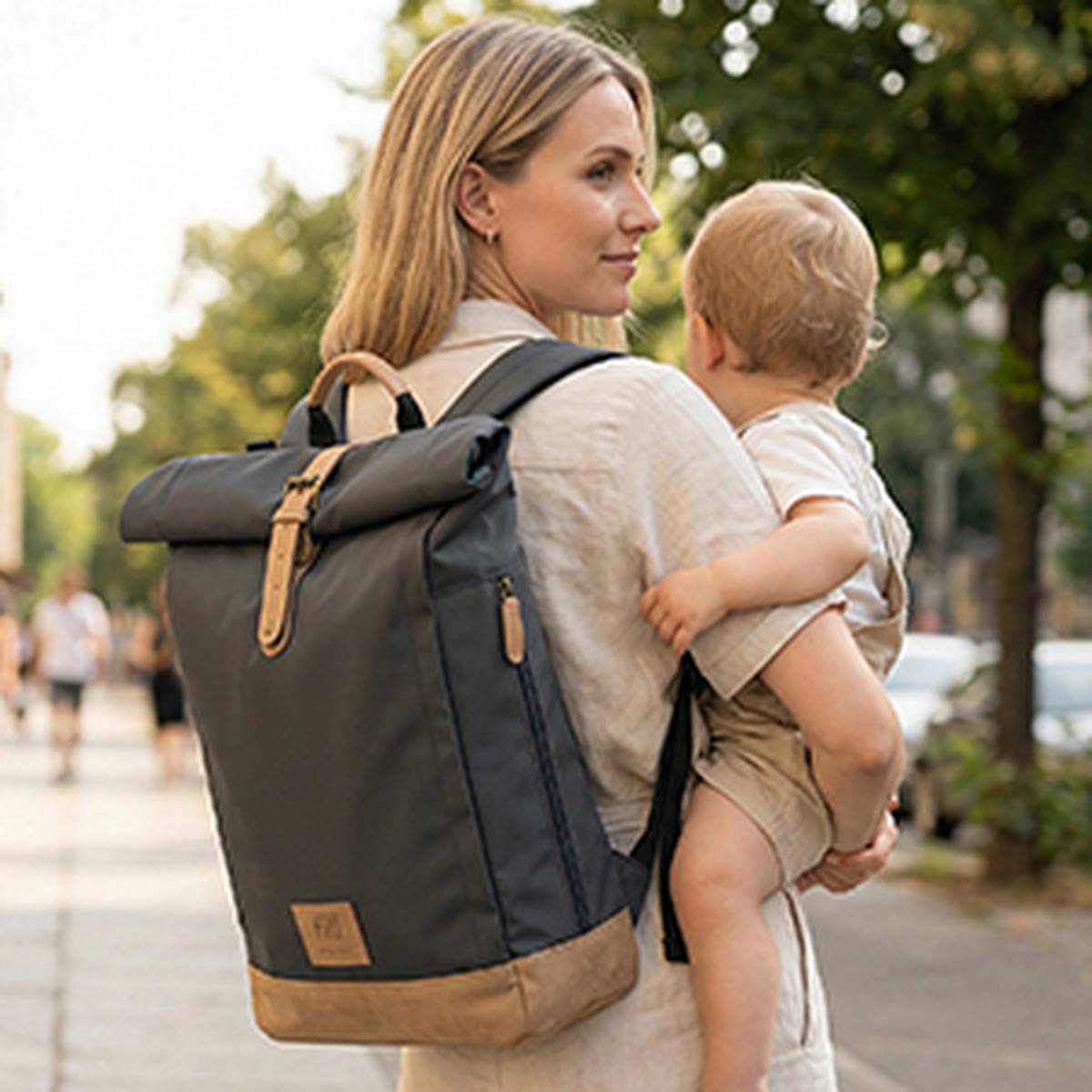 Eine Frau, die ein Baby trägt und einen großen Rucksack aufhat, geht an einem sonnigen Tag im Freien; im Hintergrund sind Bäume und Menschen verschwommen zu sehen.