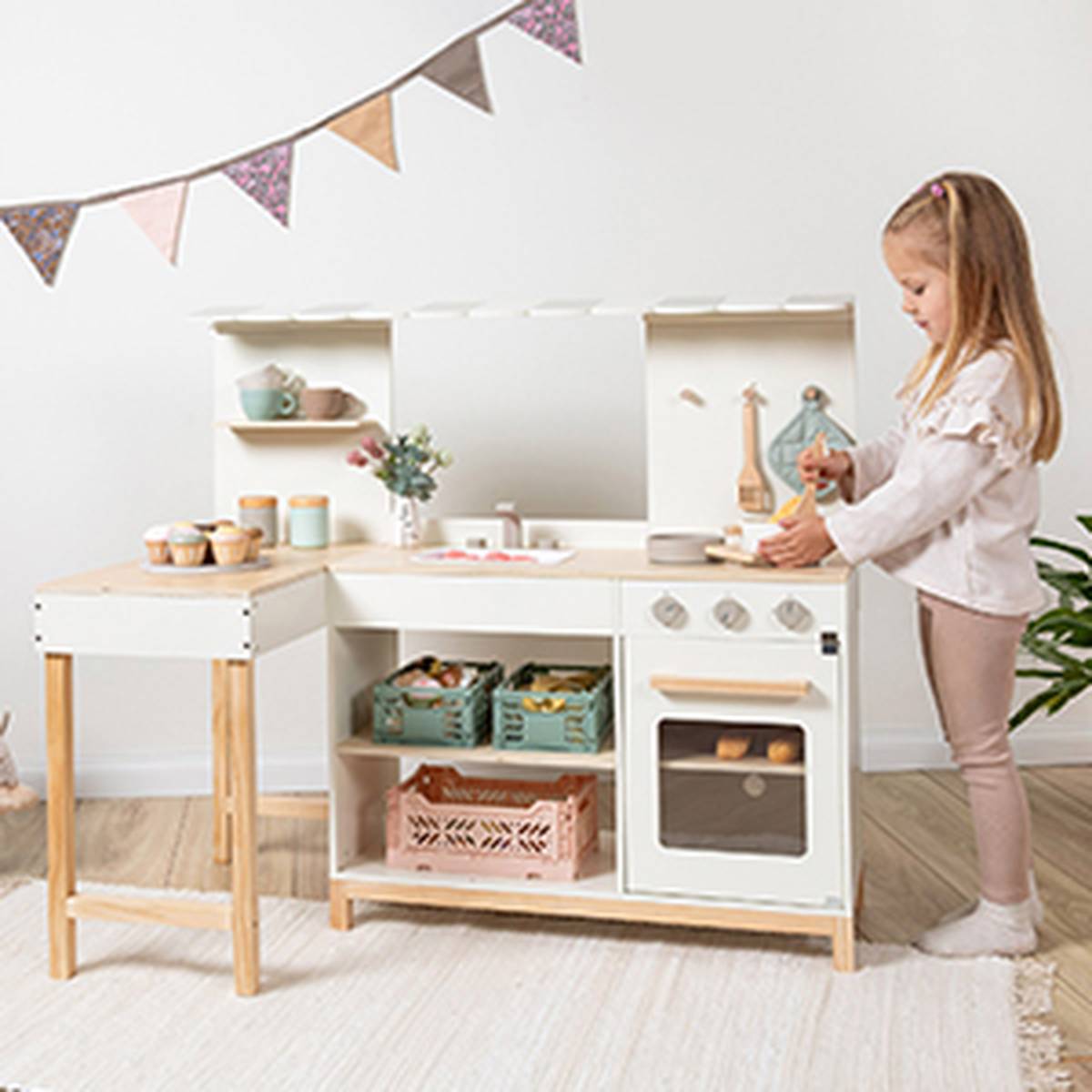 Ein kleines Mädchen spielt mit einer Spielzeugküche aus Holz – bestehend aus Herd, Backofen und Regalen – in einem mit einer Wimpelkette geschmückten Zimmer.
