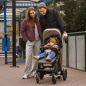 Ein Paar lächelt sein Baby im Kinderwagen an einer Bushaltestelle an; beide sind warm in Jacken und Mützen gekleidet, im Hintergrund sind Bäume zu sehen.