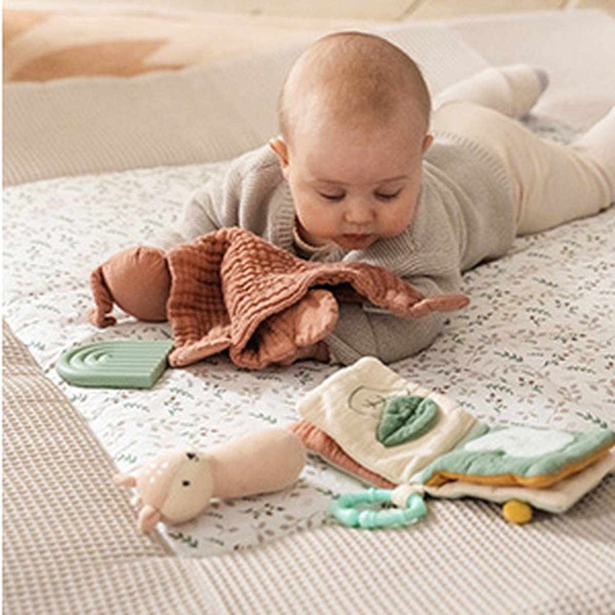 Ein Baby liegt auf einer gemusterten Decke und spielt mit Plüschtieren und einem Stoffbuch.