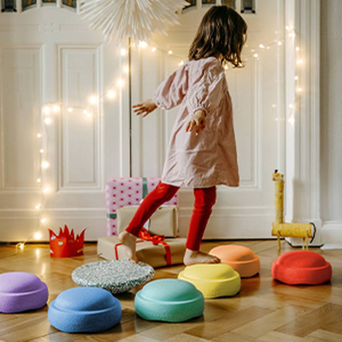 Ein Kind in einem rosa Kleid und roten Leggings balanciert auf bunten Trittsteinen in einem festlich geschmückten Zimmer mit Lichterketten und Geschenken.