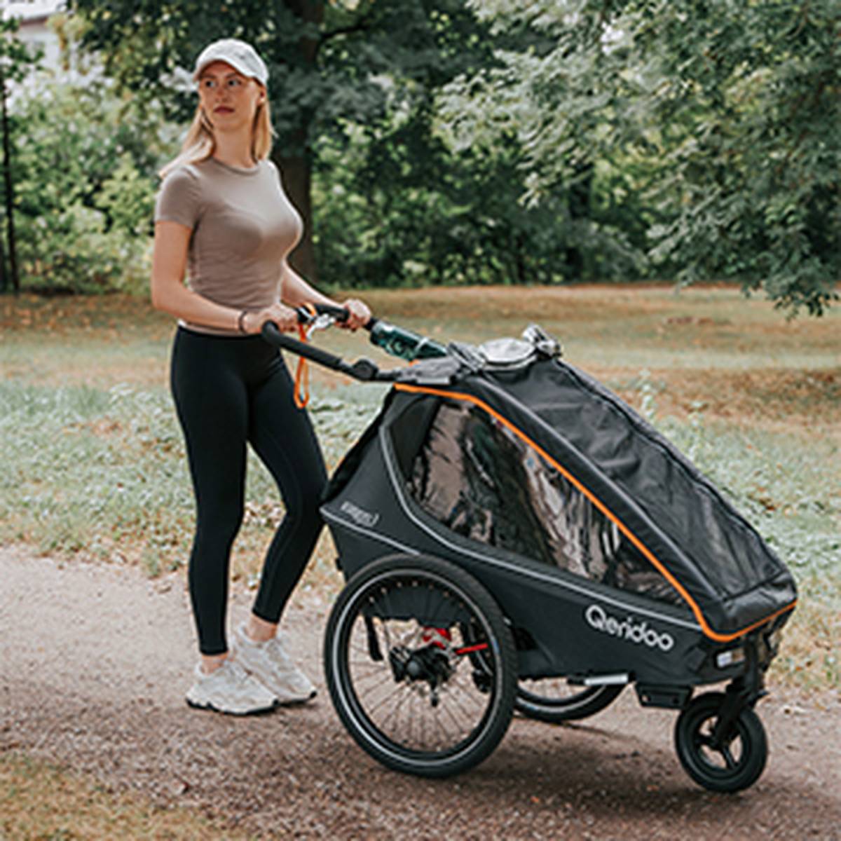 Woman in activewear pushing a three-wheeled stroller on a park path, surrounded by lush greenery.
