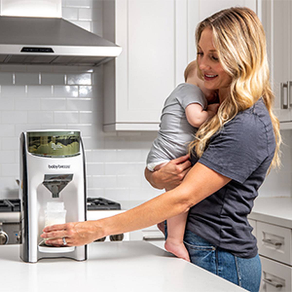 Eine Frau mit einem Baby auf dem Arm benutzt in einer modernen Küche ein Gerät von Baby Brezza und bereitet mit einer Hand ein Fläschchen zu.