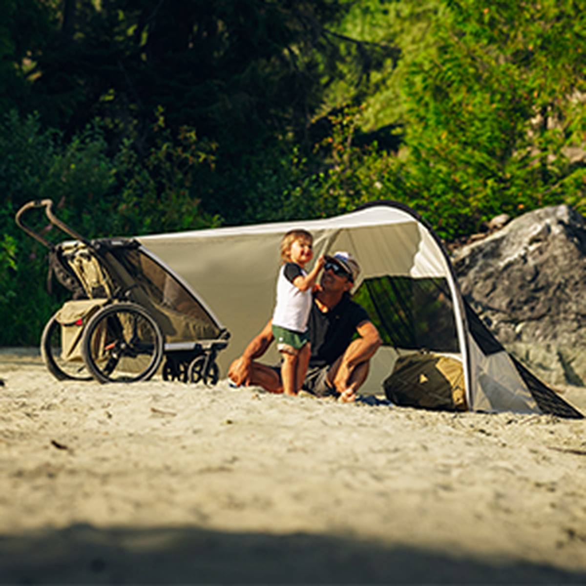 Ein Mann und ein Kind sitzen unter einem Sonnenschirm an einem Sandstrand, neben einem Kinderwagen, mit üppigem Grün im Hintergrund.