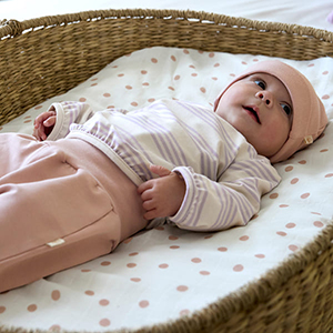 Ein Baby mit einer rosa Mütze und einem gestreiften Hemd liegt in einem Weidenkorb mit einer gepunkteten Unterlage und blickt nach oben.