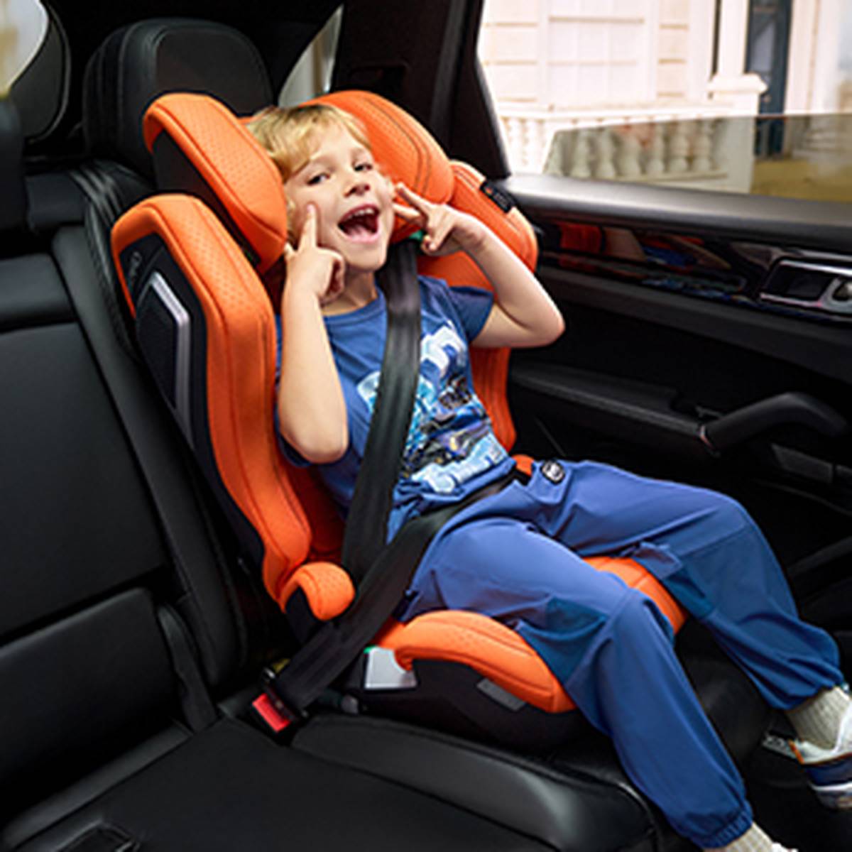 Kind in einem orangefarbenen Autositz, das lächelt und eine spielerische Geste macht, in einem blauen Outfit.