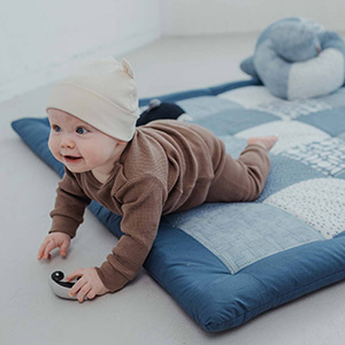Ein Baby, das ein braunes Outfit und eine cremefarbene Mütze trägt, liegt auf einer blauen Matte, hält ein kleines Spielzeug in der Hand und hat ein Plüschkissen im Hintergrund.