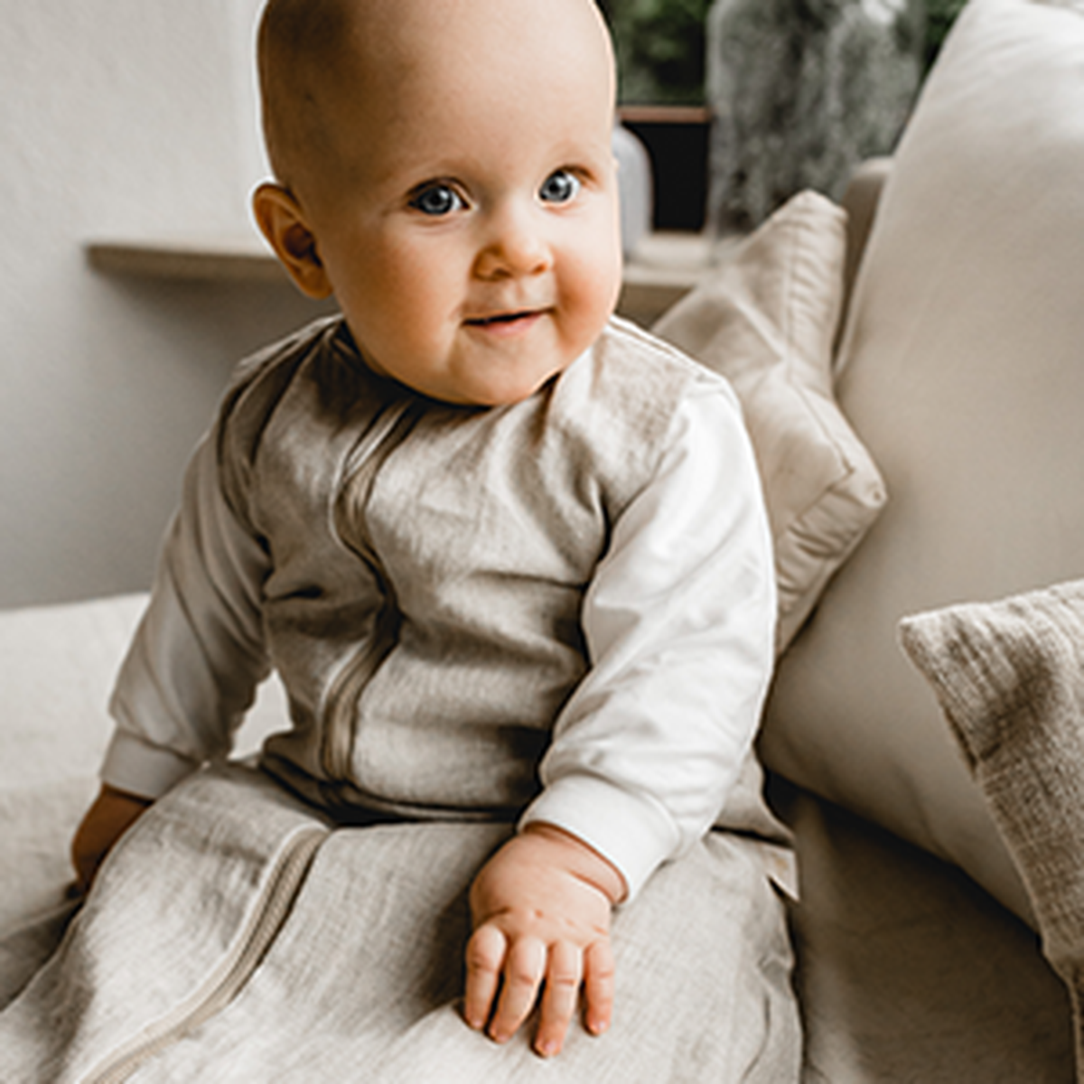 Ein lächelndes Baby in einem beigen Outfit sitzt auf einem Sofa mit weißen Kissen und blickt leicht nach oben.