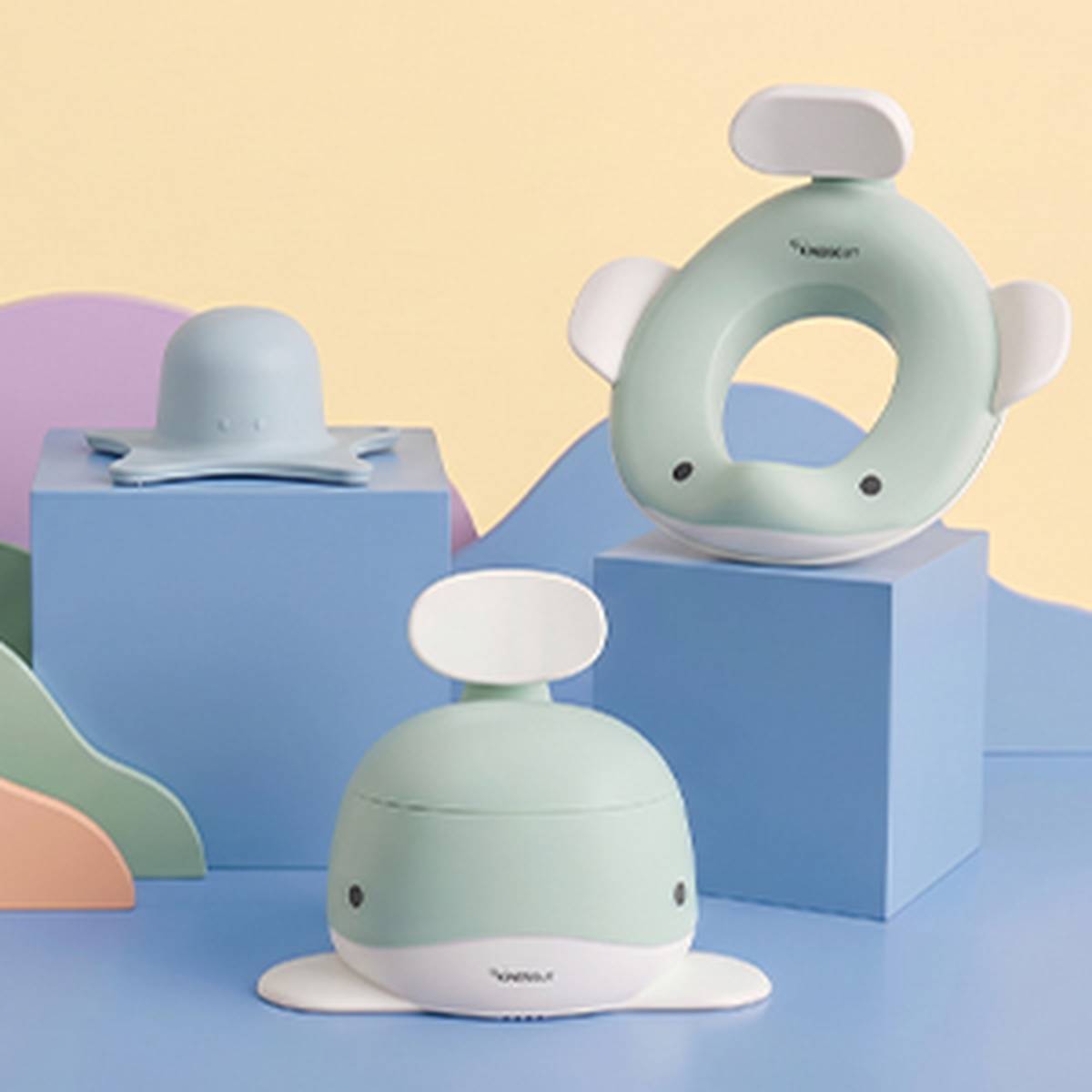 Badezubehör für Kinder in Pastellfarben, darunter ein Töpfchen mit Wal-Motiv, ein Sitzbezug und eine Sprühdüse, präsentiert auf bunten Würfelregalen.