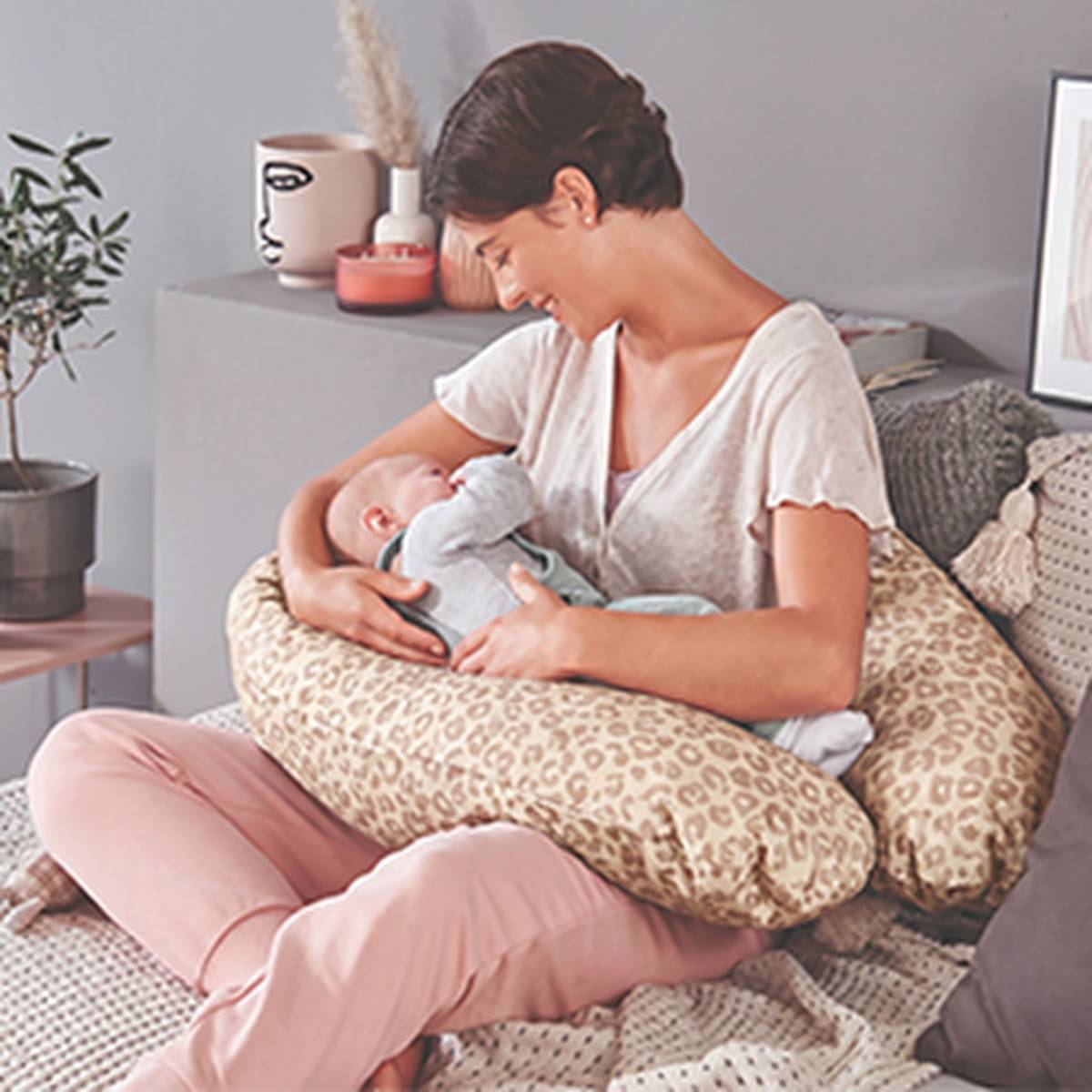 Eine Frau sitzt auf einem Bett und lächelt ein Baby an, das sie im Arm hält. Dabei verwendet sie ein Stillkissen mit Leopardenmuster. Im Hintergrund sind eine Pflanze und dekorative Wohnaccessoires zu sehen.