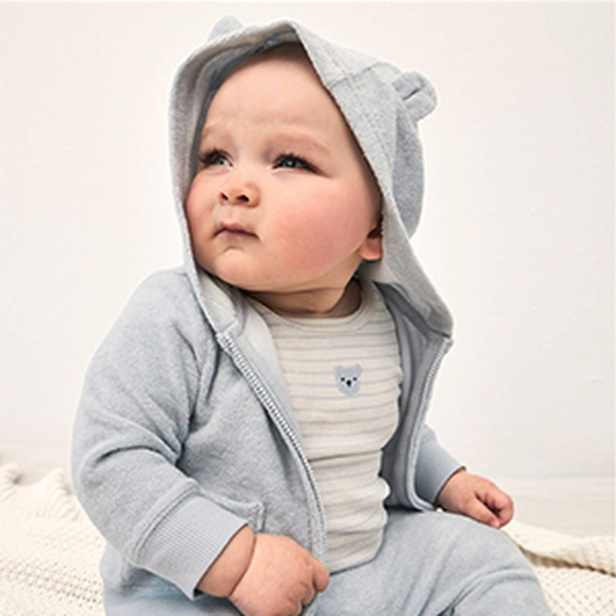 Ein Baby in einem hellblauen Kapuzenpullover mit Bärenohren blickt leicht zur Seite. Es trägt ein gestreiftes Shirt mit einem kleinen Bärenmotiv und sitzt auf einer weichen Unterlage.