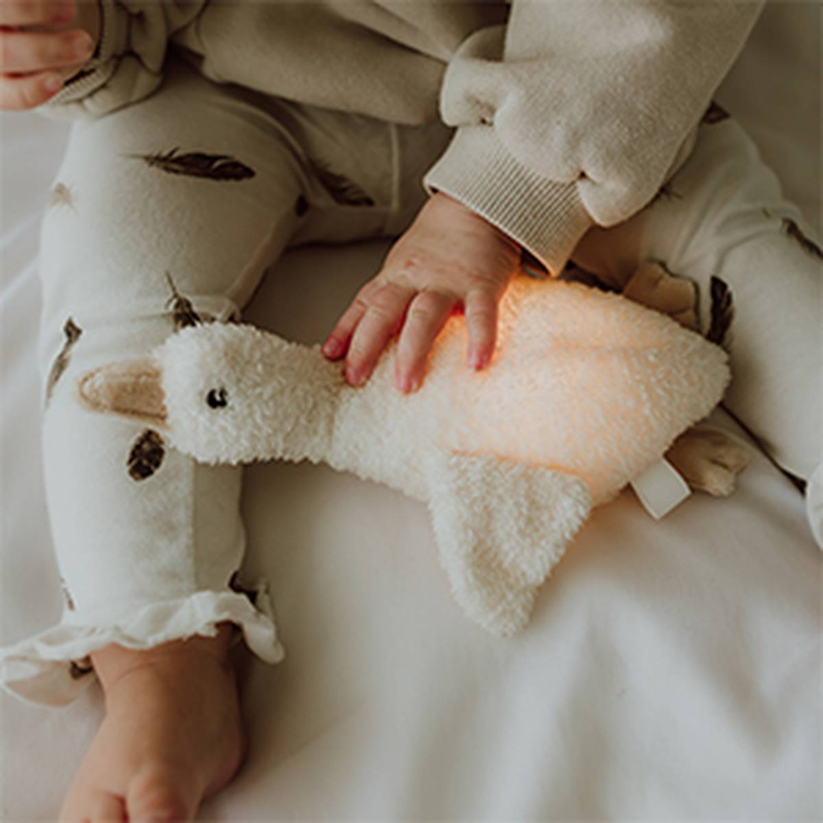 Kleine Hand berührt ein weiches Kuscheltier in Form einer Gans aus cremefarbenem Teddyfleece. Die Szene zeigt das Kuscheltier auf einer hellen Unterlage neben den Beinen einer Person in heller Kleidung und vermittelt eine gemütliche, natürliche Wohnatmosphäre.