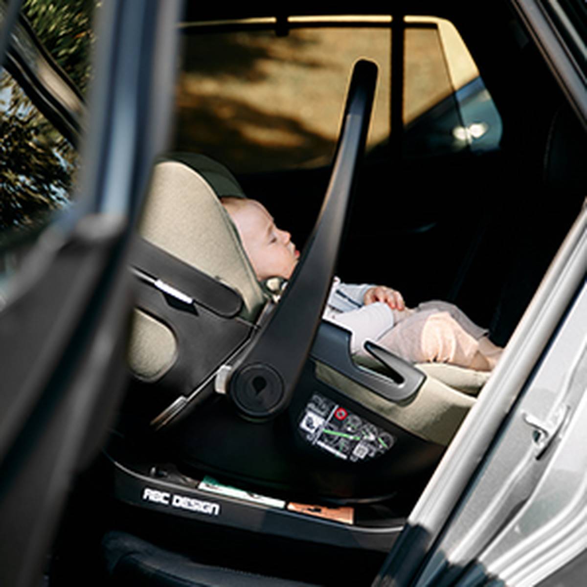 Babyschale mit Tragebügel ist auf der Rückbank eines Fahrzeugs installiert und seitlich durch die geöffnete Autotür sichtbar. Ein angeschnalltes Baby liegt in der Babyschale, die auf einer Basisstation befestigt ist.