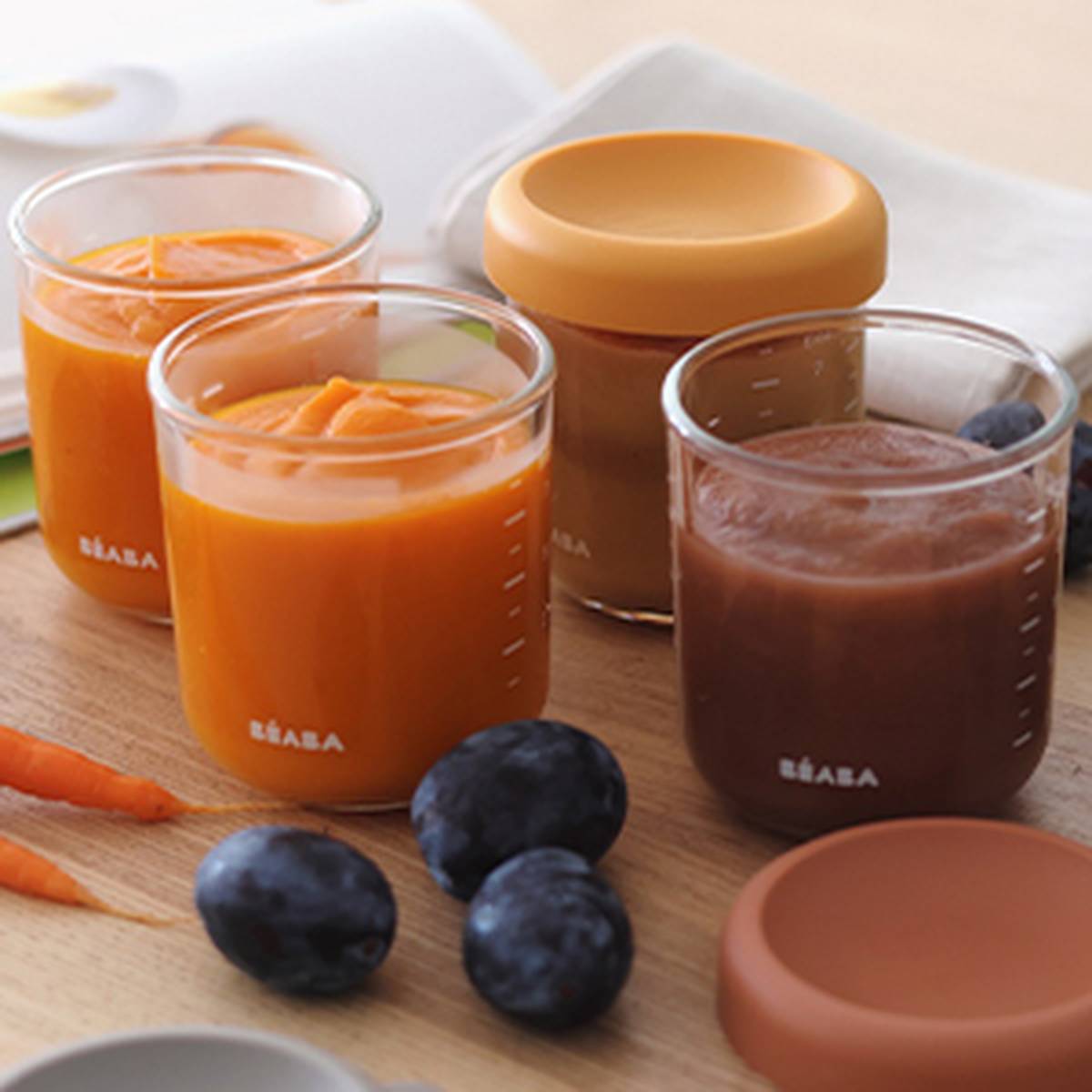 Gläser mit Babynahrung in Orange- und Brauntönen neben frischen Pflaumen und Karotten; die Deckel liegen auf einer Holzoberfläche.