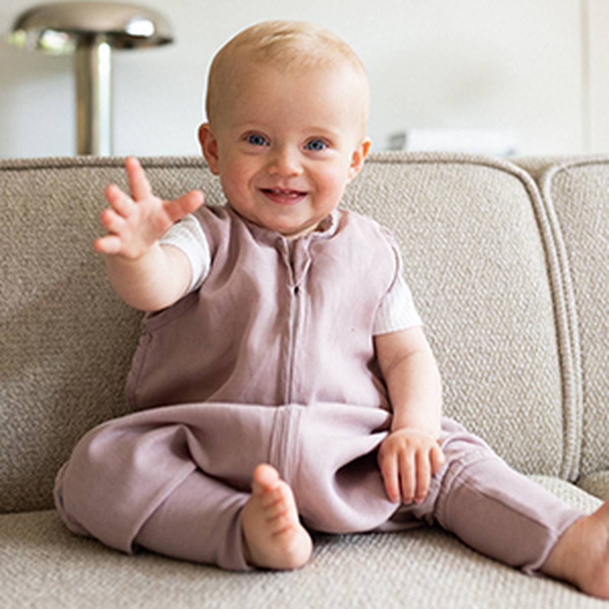 Ein lächelndes Baby in einem hellvioletten Outfit sitzt auf einem beigen Sofa, hebt eine Hand und blickt direkt in die Kamera.