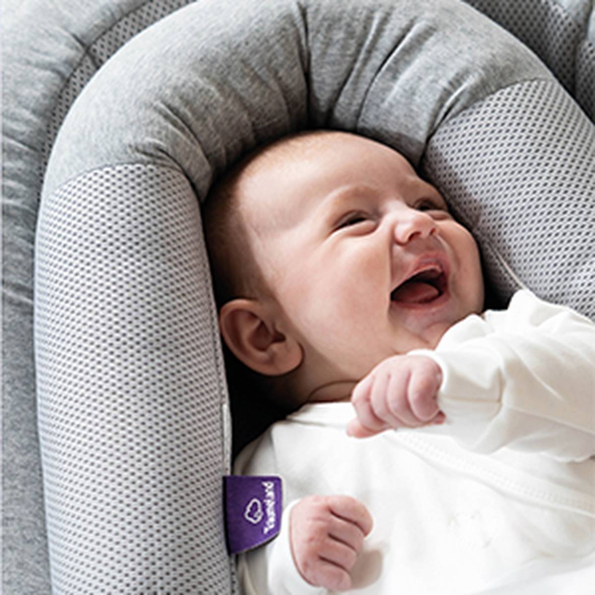 Smiling baby lying in a soft gray cushion, wearing a white outfit.