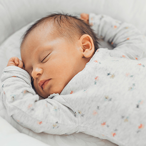 Schlafendes Baby liegt auf einer hellen Matratze oder in einem Babybett und trägt einen hellgrauen Langarm-Schlafanzug mit dezentem Sternenmuster. Die Nahaufnahme zeigt das Baby in Seitenlage vor einem weichen, hellen Hintergrund und vermittelt eine ruhige Schlafumgebung.