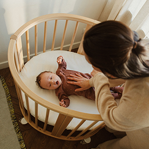 Eine Frau legt sanft ihre Hand auf die Brust eines Babys, das in einer runden Holzkrippe liegt – beide befinden sich in einem sanft beleuchteten Raum.