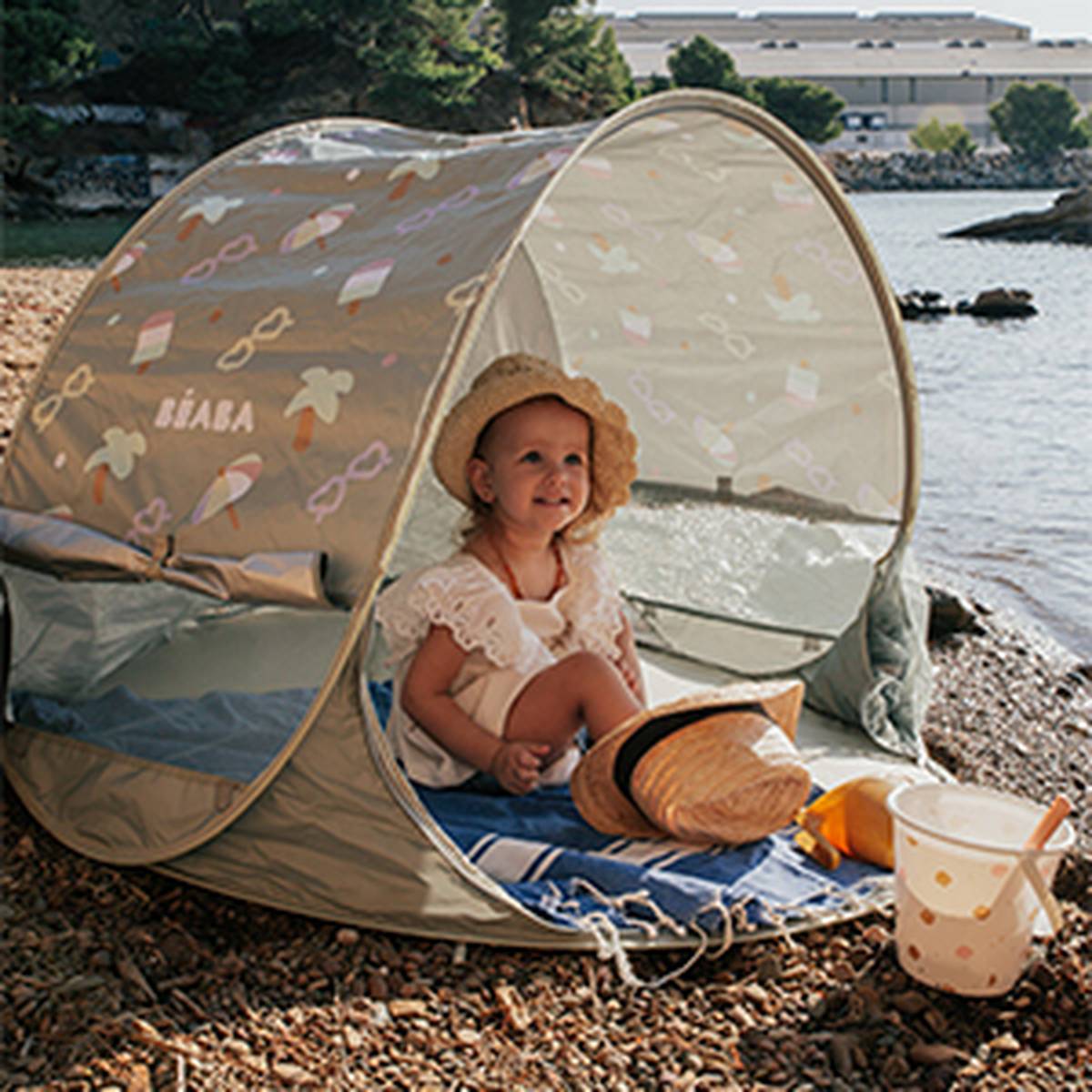 Ein Kind mit Sonnenhut sitzt in einem kleinen Strandzelt an einem felsigen Ufer; in der Nähe befinden sich ein Strohhut und ein Eimer.