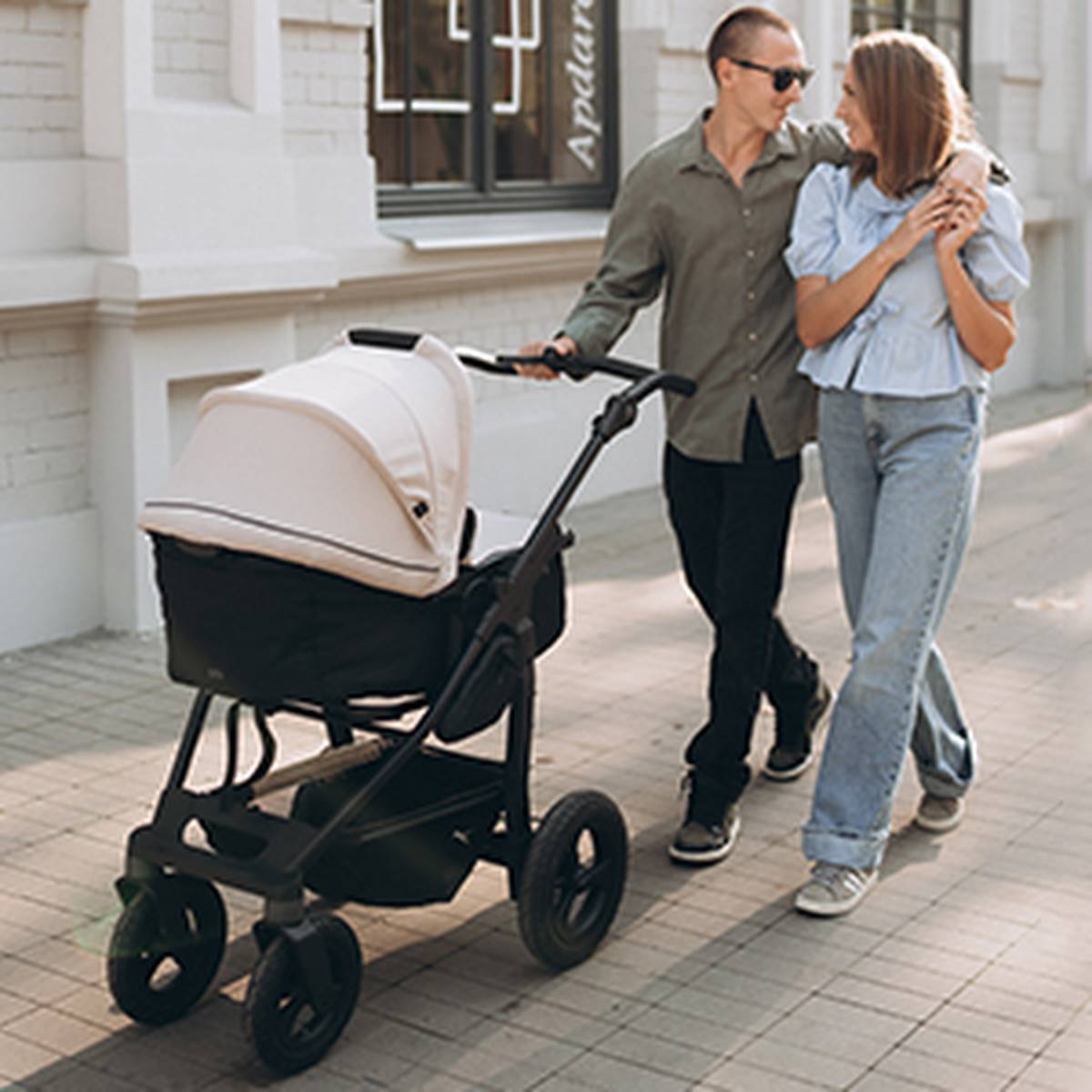 Ein Paar spaziert gemeinsam auf einem sonnigen Bürgersteig. Der Mann schiebt einen Kinderwagen, während sie sich liebevoll anlächeln.