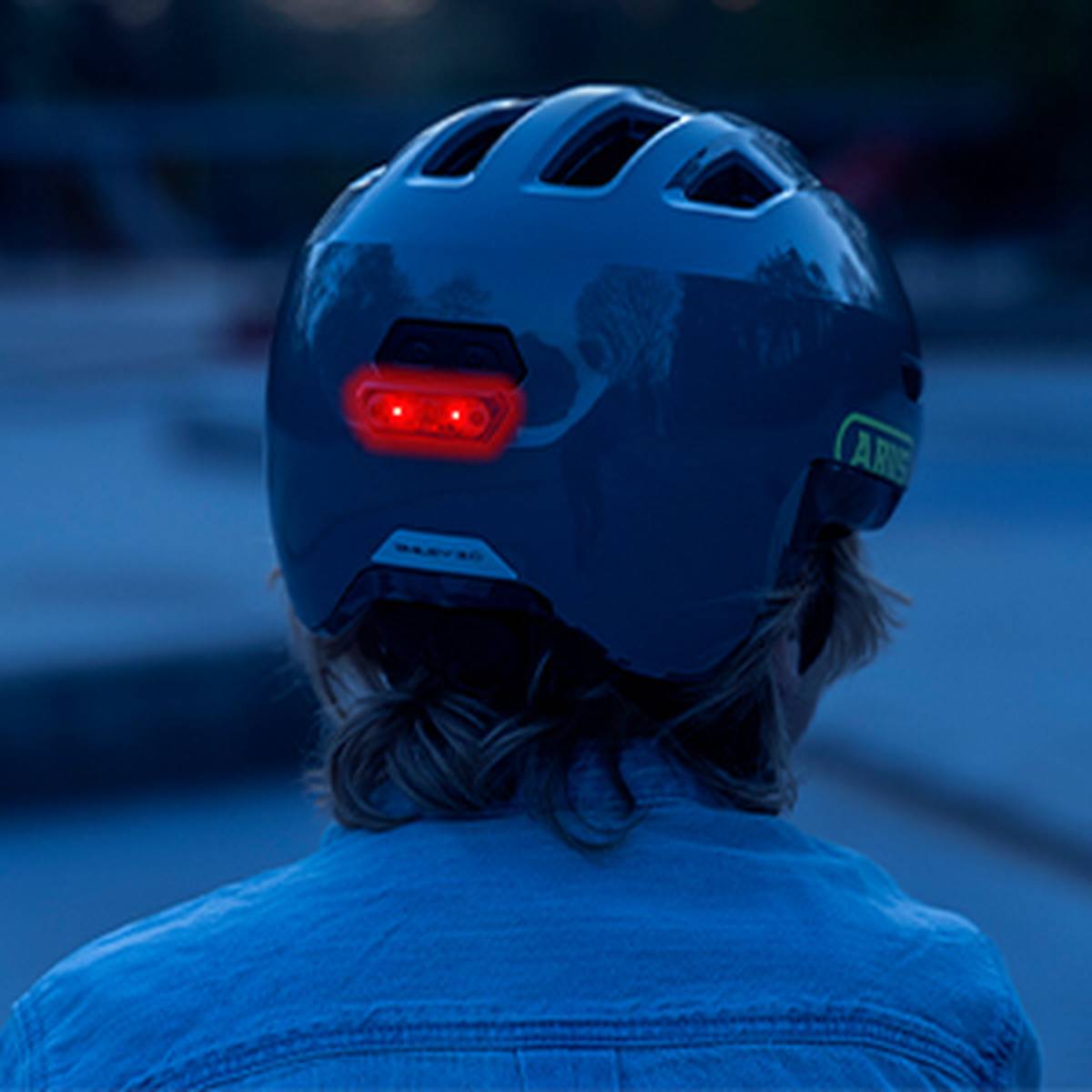 Eine Person in blauer Jacke und mit einem Helm, der an der Rückseite ein rotes Sicherheitslicht trägt; Ansicht von hinten in einem schwach beleuchteten Bereich.