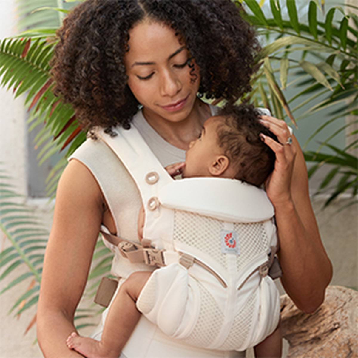 Eine Frau hält ein schlafendes Baby in einer weichen, weißen Babytrage. Sie ist von tropischen Pflanzen umgeben.