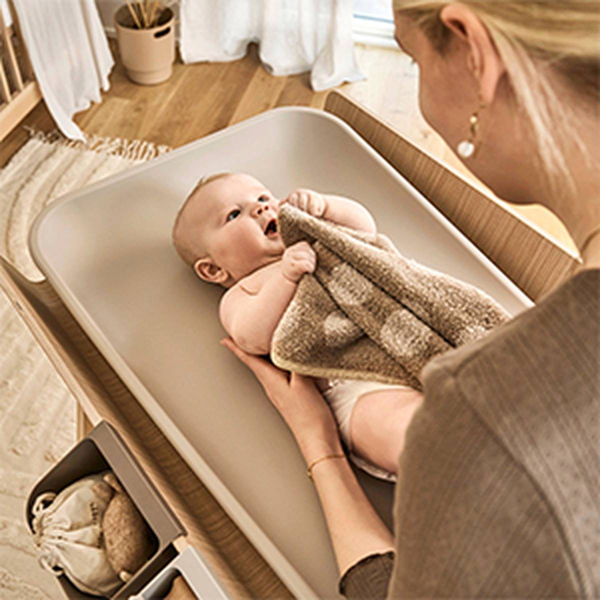 Ein Baby liegt auf einem Wickeltisch mit einer Decke in der Hand, während eine Frau in der Nähe in einem warm beleuchteten Raum steht.