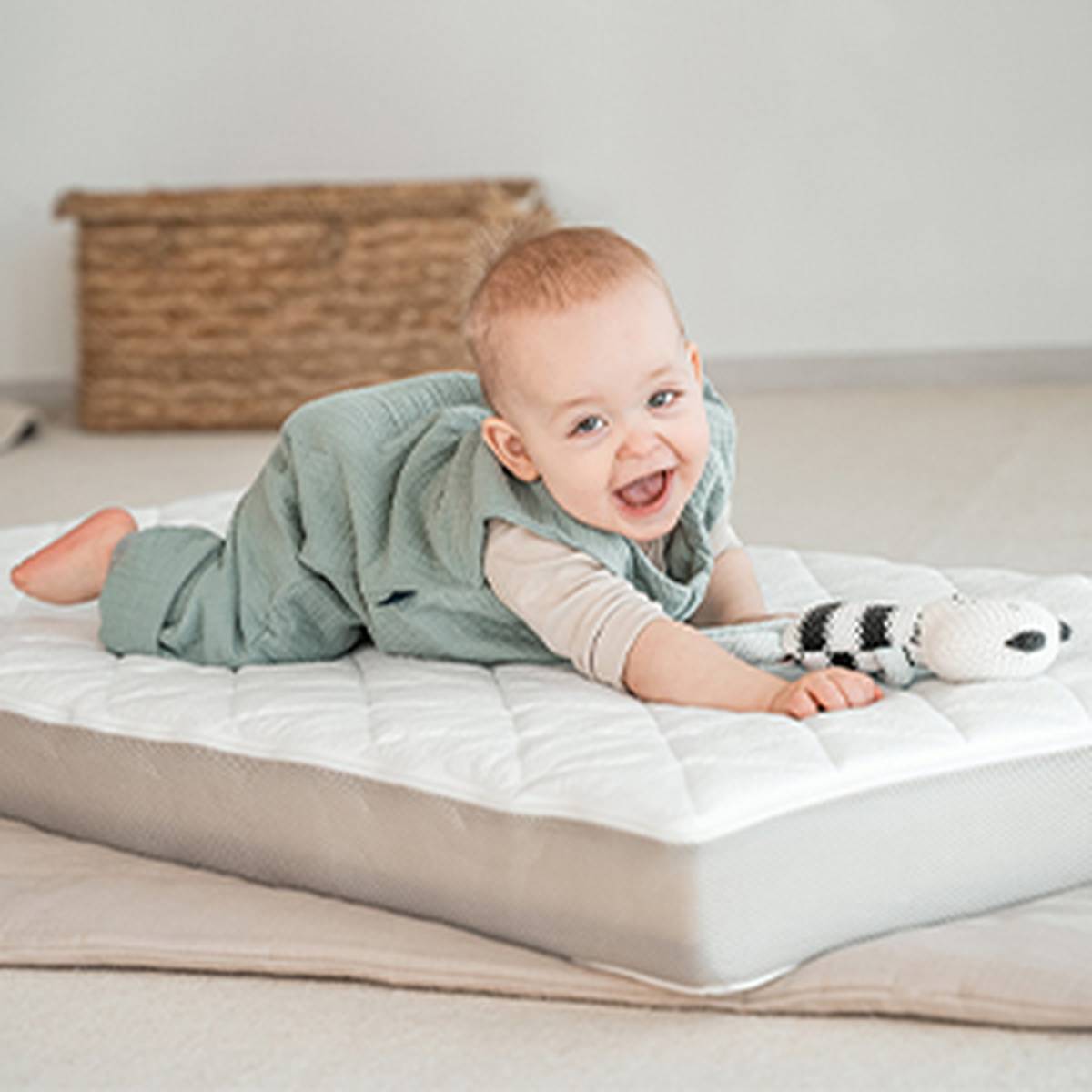 Ein lächelndes Baby in einem grünen Overall liegt auf einer Matratze und spielt in einem gemütlichen Zimmer mit einer Stoffschlange.