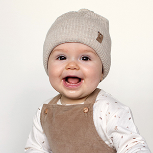Lächelndes Baby mit beigefarbener Strickmütze und brauner Latzhose über einem weißen Langarmshirt vor schlichtem Hintergrund.