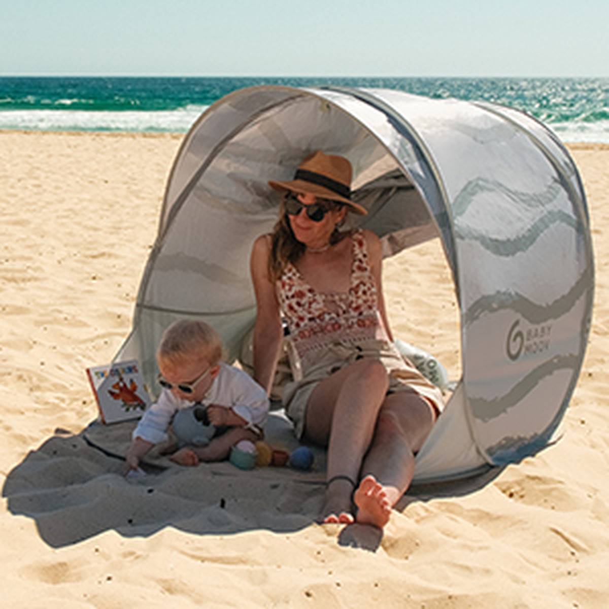Eine Frau und ein Baby sitzen unter einem mobilen Sonnenschirm an einem Sandstrand; beide tragen Sonnenbrillen. Im Hintergrund sind strahlender Sonnenschein und das Meer zu sehen.