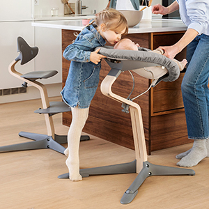 Ein junges Mädchen küsst ein Baby, das in einem Hochstuhl sitzt, während in einer modernen Küche ein Erwachsener daneben steht.