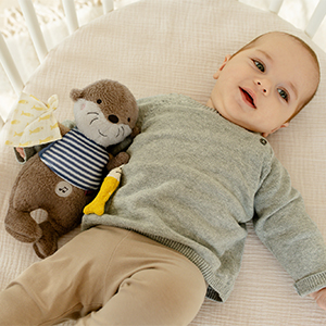 Ein Baby in einem grauen Pullover liegt in einem Gitterbett und hält einen Plüsch-Otter mit gestreiftem Hemd im Arm.