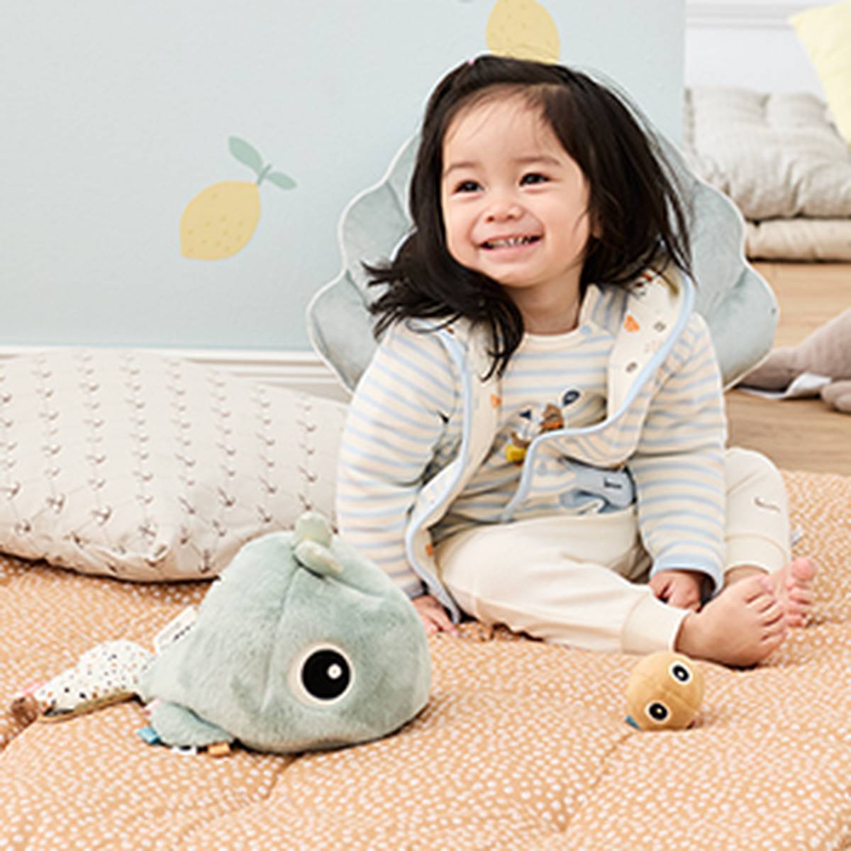 Ein lächelndes Kleinkind sitzt in einer gemütlichen Zimmerumgebung auf einer gemusterten Matte mit Stofftieren. Es trägt eine gestreifte Jacke und eine cremefarbene Hose.