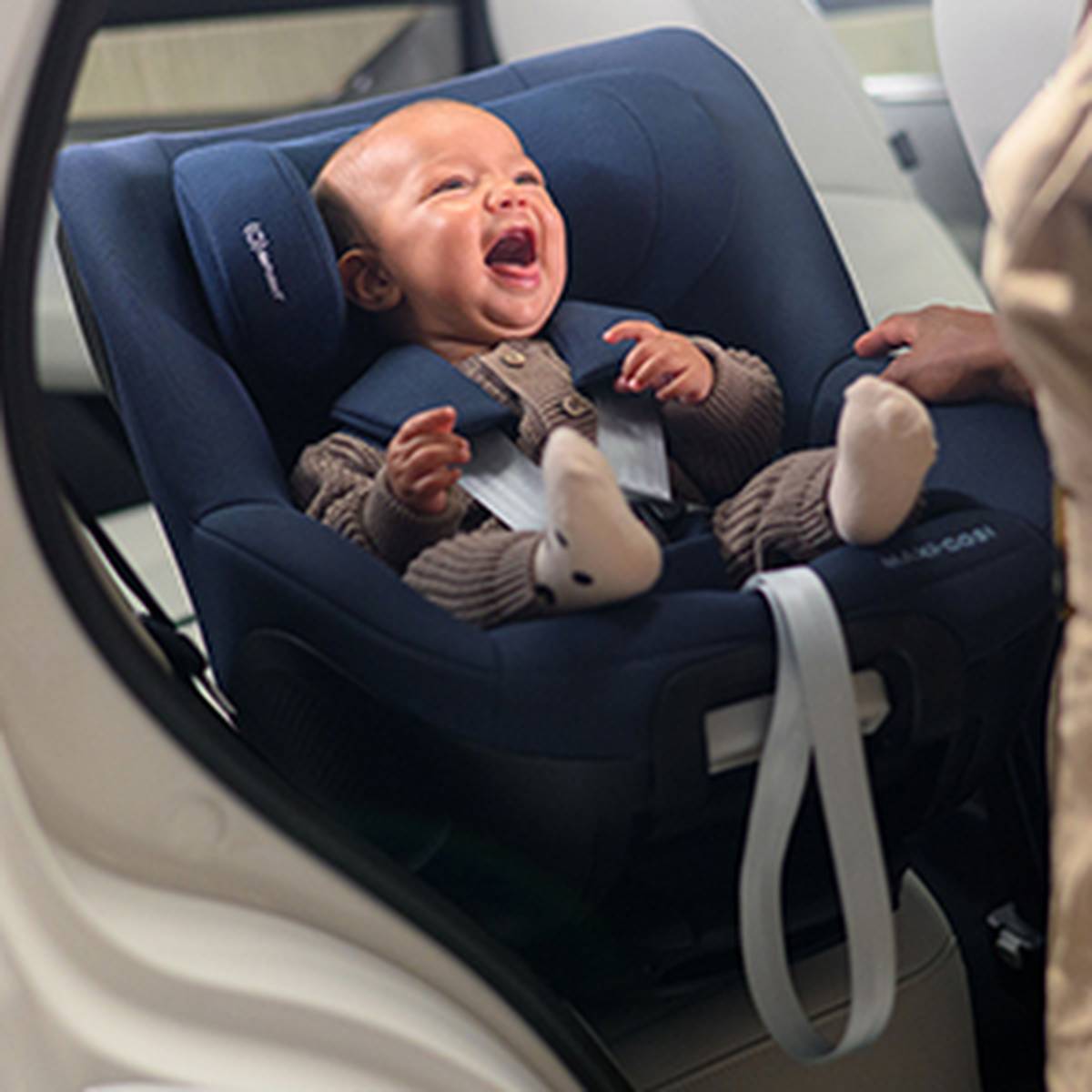 Ein lächelndes Baby in brauner Kleidung sitzt sicher in einem dunkelblauen Kindersitz, der auf der Rückbank eines Fahrzeugs montiert ist. Ein Erwachsener hält die Hand des Babys.