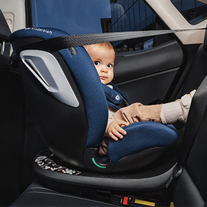 Ein Baby sitzt in einem blauen Kindersitz in einem Fahrzeug, ist mit Sicherheitsgurten gesichert und blickt in die Kamera.