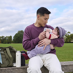 Ein Mann in einem lila Pullover füttert ein Baby, das eine Mütze trägt, mit der Flasche; er sitzt dabei auf einer Parkbank. Neben ihm stehen eine Tasche und eine Thermoskanne.