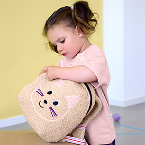 Kind mit rosafarbenem T-Shirt hält einen flauschigen Rucksack in Katzenform von LÄSSIG vor hellem Innenraumhintergrund.