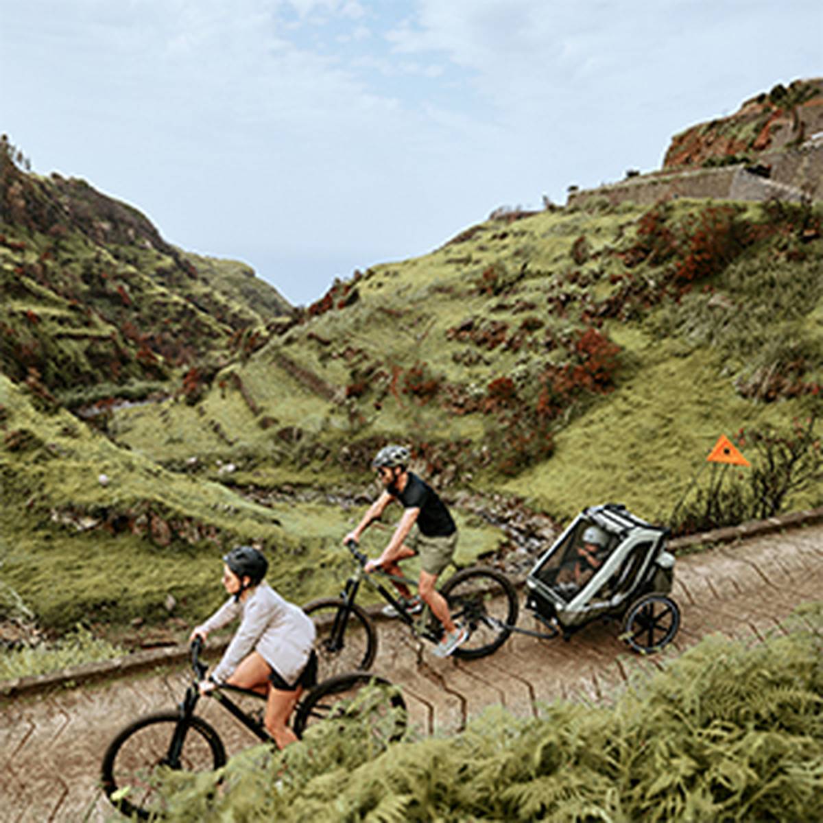 Zwei Radfahrer fahren auf einem malerischen Bergweg und ziehen ein Kind in einem Fahrradanhänger hinter sich her. Die Landschaft ist geprägt von üppig grünen Hügeln und einem bewölkten Himmel.