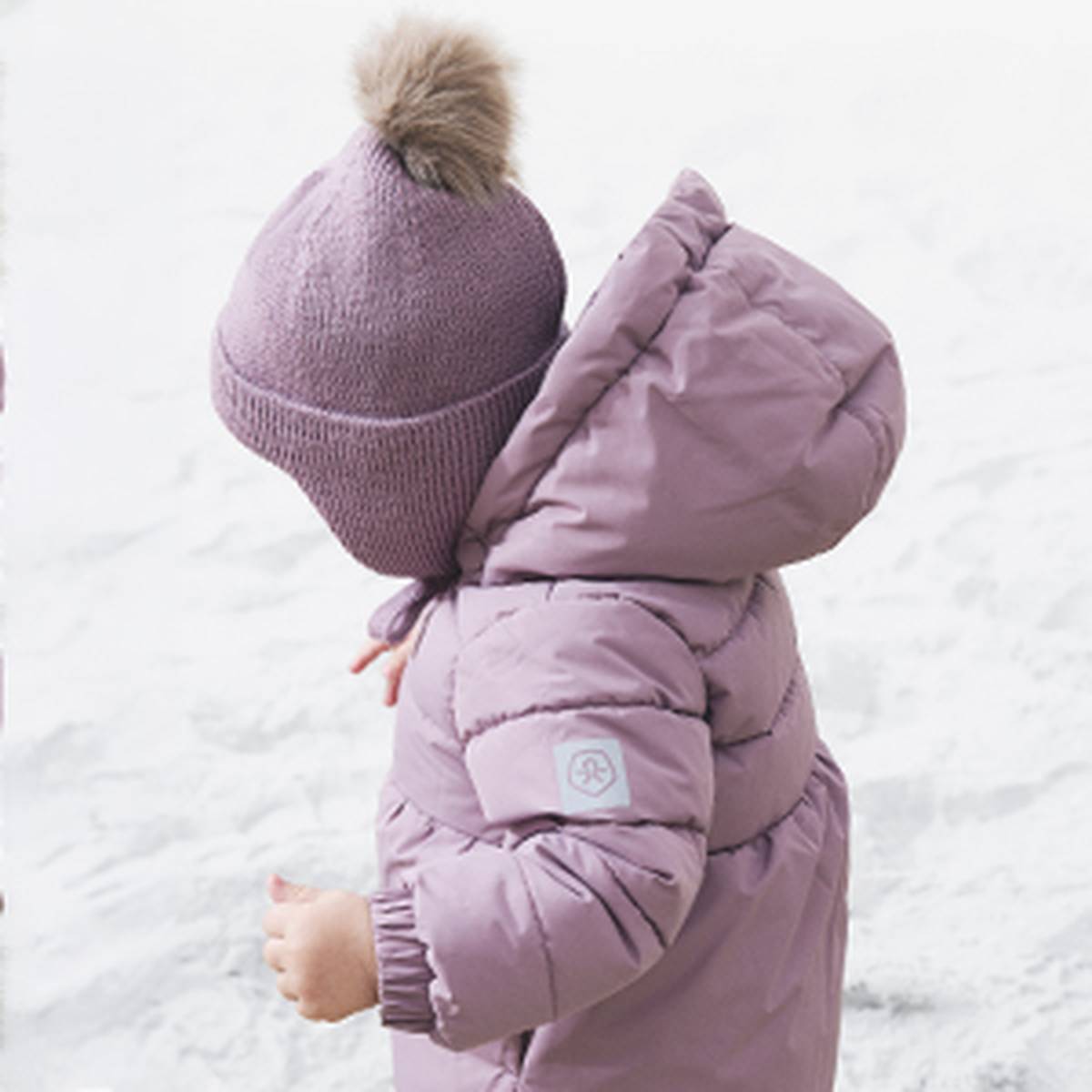 Ein Kind in einem lavendelfarbenen Mantel und mit einer Mütze mit Bommel steht auf verschneitem Boden und blickt vom Betrachter weg.