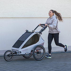 Eine Frau joggt mit einem dreirädrigen Jogging-Kinderwagen vor einer hellen Wand; sie trägt einen Kapuzenpullover, Leggings und Turnschuhe.