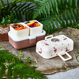 Bento-Box mit zwei offenen Fächern, gefüllt mit Essen, vor einem natürlichen Hintergrund mit Grünpflanzen und Farnen. Der Deckel der Box ist mit Fuchsmotiven verziert.