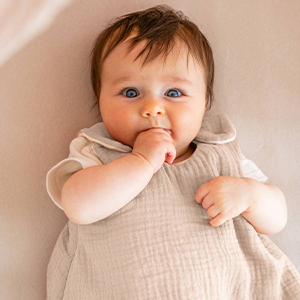 Ein Baby liegt auf einer weichen Unterlage, trägt helle Kleidung, hat eine Hand im Mund und blickt mit großen Augen nach oben.