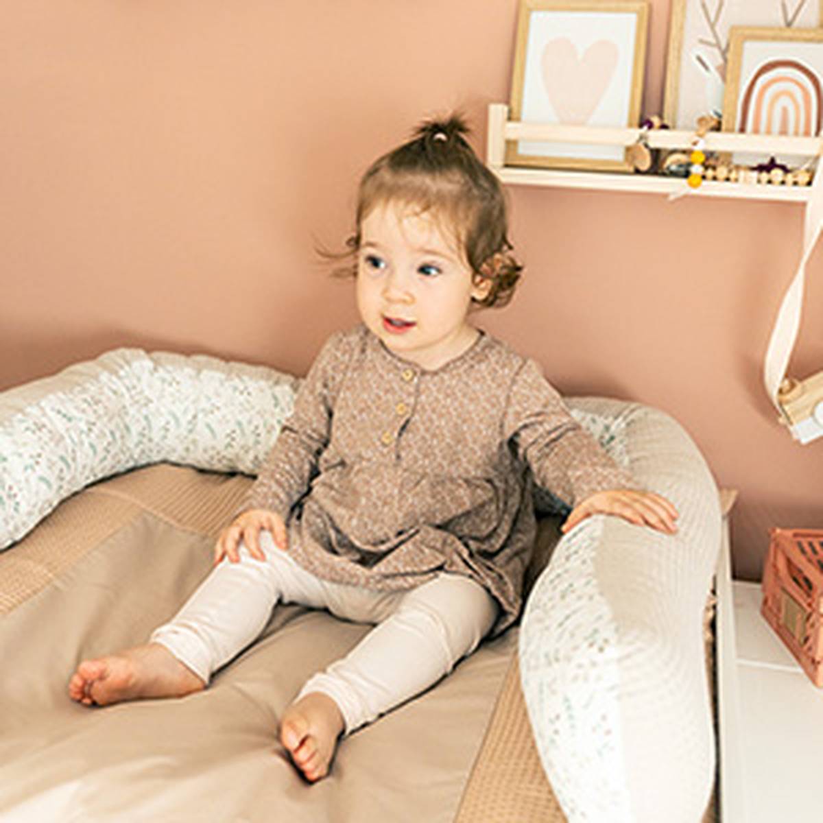 Ein Kleinkind sitzt auf einem gepolsterten Bett in einem Kinderzimmer in warmen Farbtönen, trägt ein gemustertes Kleid und Leggings und ist von Dekoartikeln und Spielzeug umgeben.