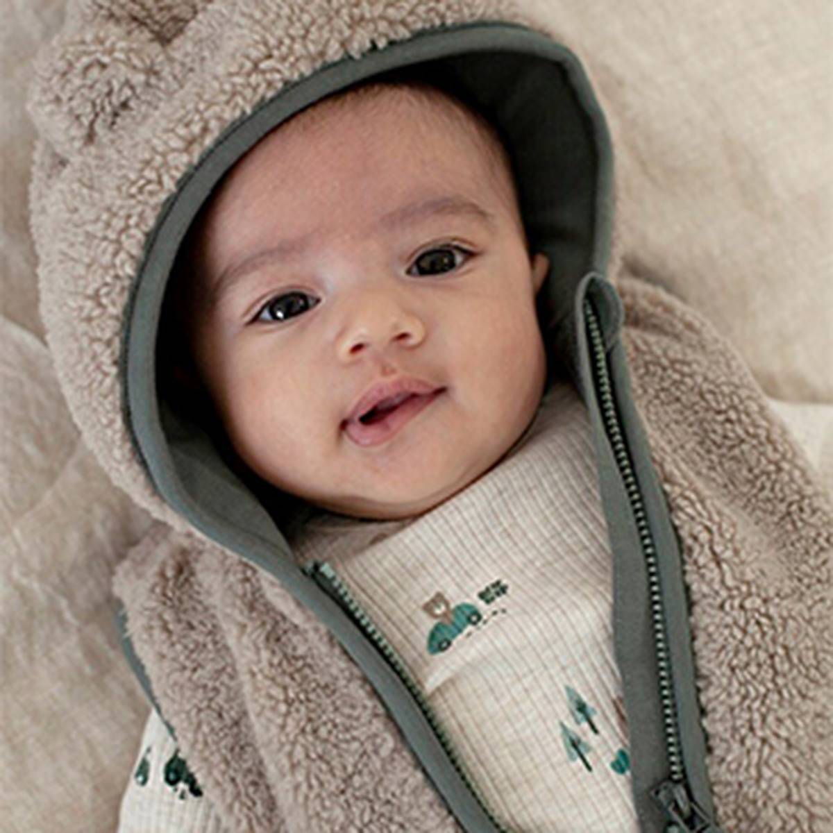 A baby wearing a cozy hooded outfit with bear ears, smiling softly while wrapped in a fluffy blanket.