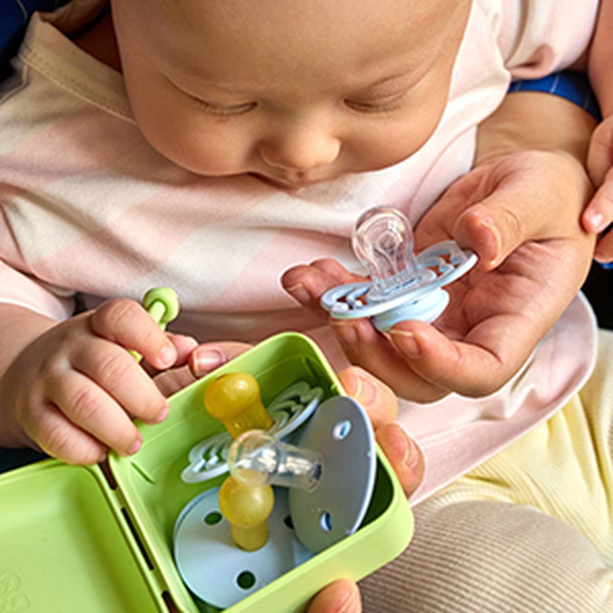 Ein Baby mit einem Schnuller im Mund, auf dessen Schoß ein offenes grünes Etui mit weiteren Schnullern liegt, während es gemütlich auf dem Arm gehalten wird.