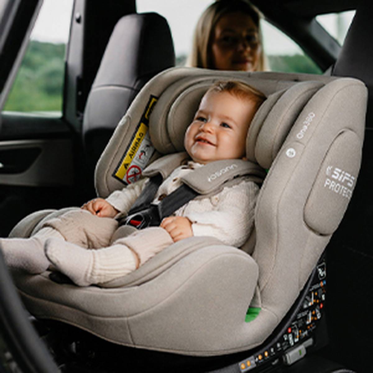 Ein lächelndes Baby sitzt sicher in einem grauen Autositz in einem Fahrzeug, während sich im Hintergrund eine erwachsene Person in der Nähe befindet.